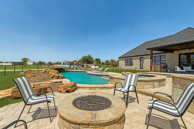 Fire pit with chairs on a stone patio, with the pool and spa behind it